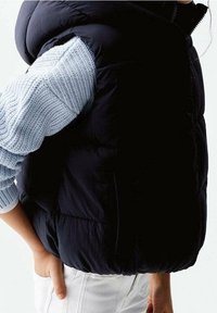 Navy puffer vest with a quilted design, paired with a light blue textured knitted sweater sleeve. White pants and a relaxed fit.