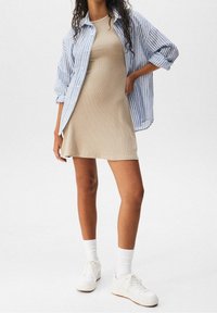 Abito a coste beige con maniche corte, indossato sotto una camicia a righe blu e bianche con bottoni. Abbinato a sneakers bianche e calze alla caviglia.