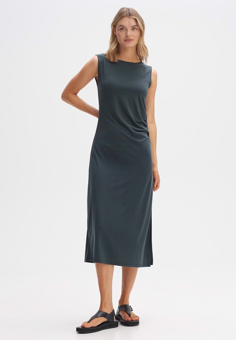 Ärmelloses, dunkelgrünes Midikleid mit entspanntem Schnitt, seitlichen Schlitzen und glatter Textur. Kombiniert mit flachen schwarzen Sandalen.