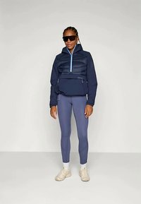 Jūras spēku pūkains jaka ar gaiši zilu rāvējslēdzēju, priekšējo kabatu; apvienota ar pieguļošām, gaiši zilām legingām un bēšām kedām. Modelis nēsā saulesbrilles.