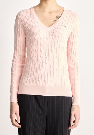 Femme portant un pull en maille torsadée rose clair à col en V et un pantalon noir à fines rayures, montrant des tatouages floraux sur la clavicule et le poignet.