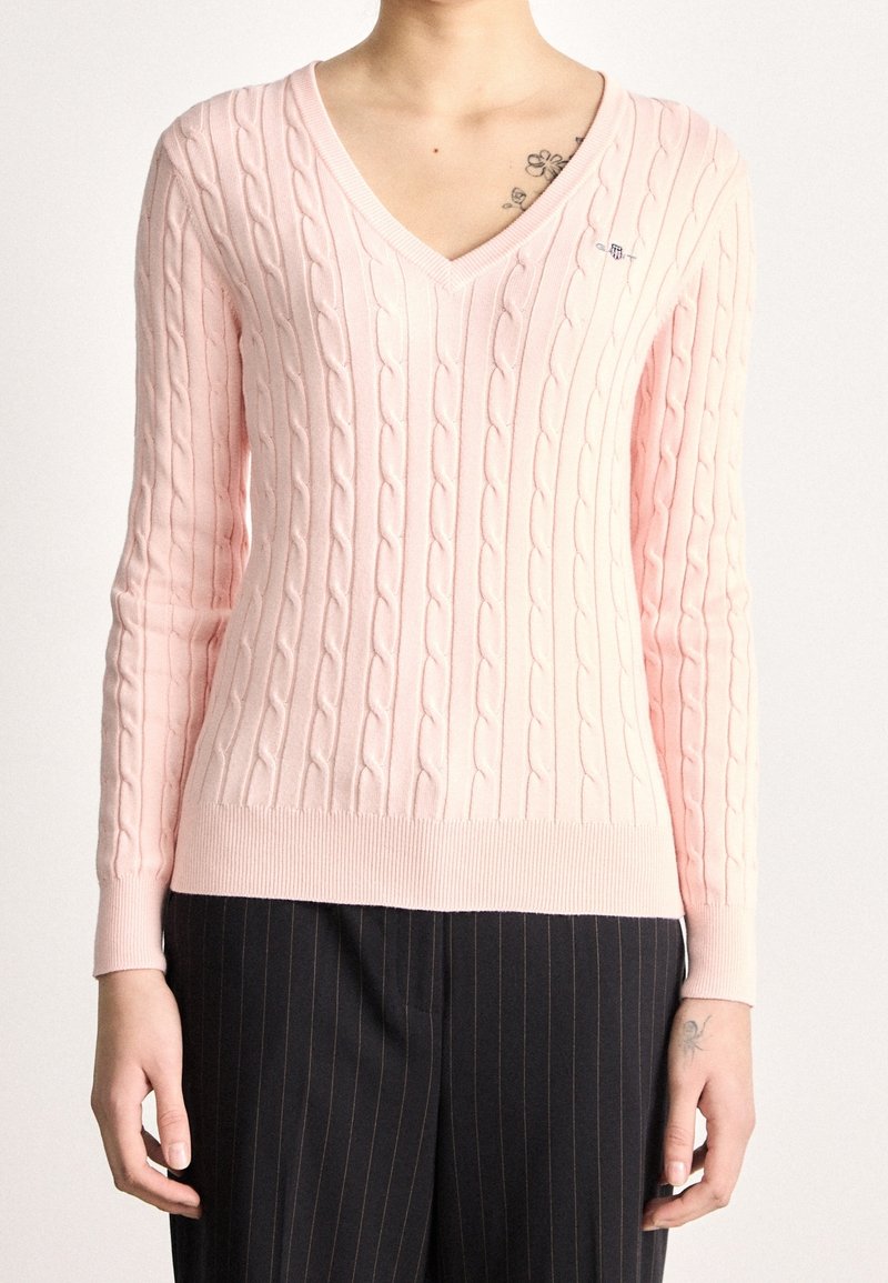 Femme portant un pull en maille torsadée rose clair à col en V et un pantalon noir à fines rayures, montrant des tatouages floraux sur la clavicule et le poignet.
