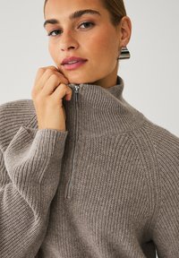 Brun strikket pullover med høj krave og en halv lynlås. Har ribbet detaljering på kraven og manchetterne. Glat tekstur.