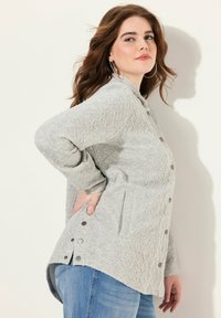 Světle šedý texturovaný svetr s diamantovým vzorem, cvakací knoflíky na bocích a dvěma předními kapsami. Dlouhé rukávy, uvolněný střih.