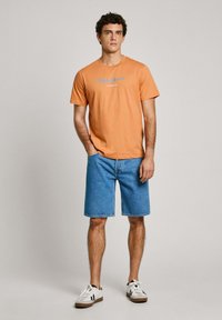 Oranžinė medvilninė marškinė su mėlynu logotipu, derinamos su mėlynomis džinsinėmis šortais. Modelis, dėvintis baltus sportbačius su juodais akcentais, stovi neutraliame fone.