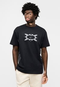 Μαύρο βαμβακερό μπλουζάκι με κεντρικό γκρι λογότυπο Nike και περίτεχνα μοτίβα στο πλάι. Ο σχεδιασμός είναι σε χαλαρή εφαρμογή με κοντά μανίκια.