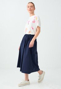 Jeune femme debout portant un haut blanc à motifs floraux, une longue jupe bleu marine et des baskets blanches sur un fond clair uni.