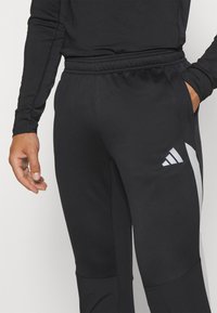 Schwarze Sporthosen aus strukturiertem Stoff, mit einem weißen Adidas-Logo an der linken Hüfte und seitlichen Taschen zur Aufbewahrung.