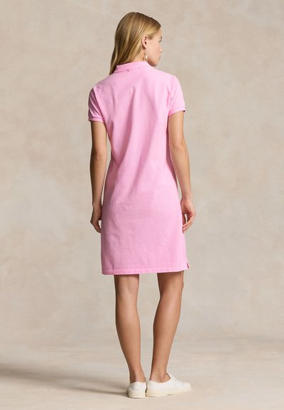 Woman standing, facing away, wearing a knee-length pink polo dress and white sneakers against a neutral background.