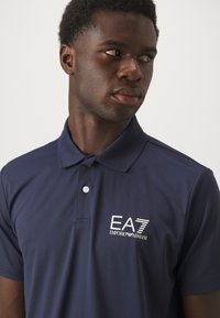 Navy blue polo shirt made of breathable fabric, features a two-button placket and embroidered EA7 logo on the left chest.