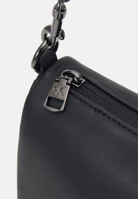 Černá taška z umělé kůže se zipovým uzávěrem a kovovým přívěskem na zip v podobě loga CK. Doplněná lesklým černým řetízkovým popruhem.