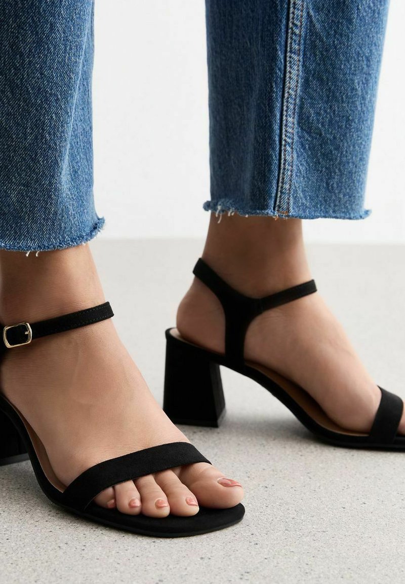 Sandalias de tacón de ante negro con tacón de bloque, banda ajustable en el tobillo y diseño de punta abierta. Llevadas con jeans de mezclilla recortados que presentan dobladillos deshilachados.