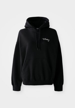 Črna majica s kapuco, sprednjim žepom in vrvicami. Ima belo vezeno besedilo "obey" na prsih. Mehka tekstura tkanine.