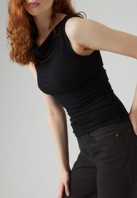 Juodas rankovių viršus su drapiruotu iškirpte ir surinktais šonais, derinamas su tamsiais džinsais. Audinys atrodo lygus ir tamprus.