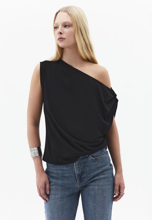 Femme blonde portant un top noir une épaule et un jean bleu, avec un bracelet manchette argenté à son poignet gauche, debout sur fond blanc.