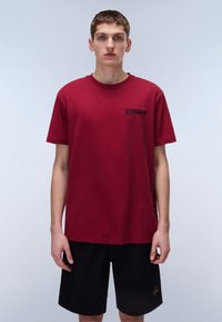 Bordeaux-rotes Baumwoll-T-Shirt mit kurzen Ärmeln, rundem Ausschnitt und einem kleinen schwarzen Logo auf der linken Brust. Getragen über schwarzen Shorts.