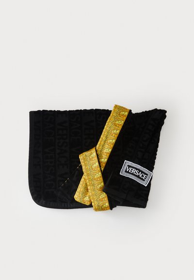 Fekete bársony borítású kézitáska, amelyen dombornyomott "Versace" logó látható, arany mintázatú pánttal és fekete háttéren fehér feliratú címkével.