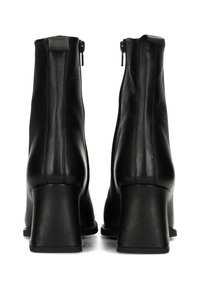 Par de botines negros de cuero con tacones gruesos y cremalleras traseras, mostrados desde atrás sobre un fondo blanco.