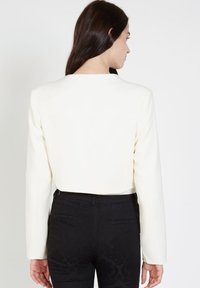 Blazer blanco recortado con escote cuadrado y mangas largas, combinado con pantalones negros texturizados. Tejido suave; detalles mínimos.