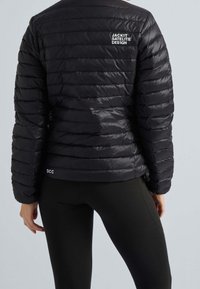 Personne portant une veste matelassée noire avec du texte blanc au dos et un legging noir, debout devant un fond gris uni.