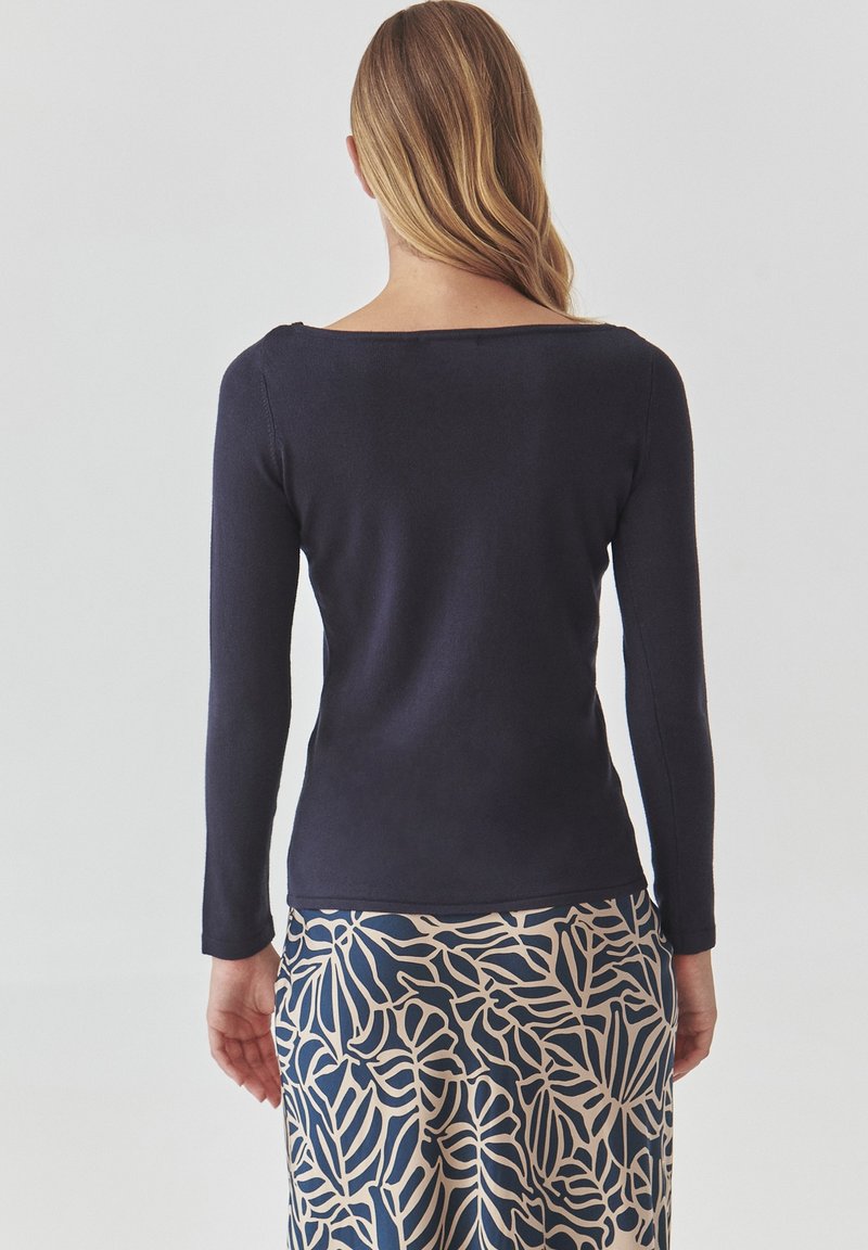 Granatowa bluzka z długim rękawem i szerokim dekoltem, zestawiona ze spódnicą w jasny wzór botaniczny na ciemnym tle.