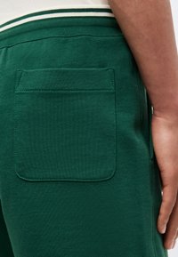 Κοντινό σεγκόνατου ενός ατόμου που φοράει πράσινη φόρμα με πίσω τσέπη με κάλυμμα και λευκή μέση με πράσινες ρίγες.