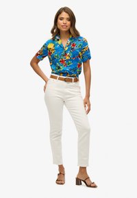 Camisa corta de manga floral azul con acentos amarillos y verdes, combinada con pantalones blancos ajustados y sandalias de taco marrones. Detalle de cinturón marrón visible.
