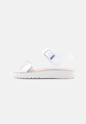 Weiße flache Sandale mit gepolsterter Sohle, verstellbarem Knöchelriemen mit silberner Schnalle und funkelnden, gekreuzten Vorderriemen.
