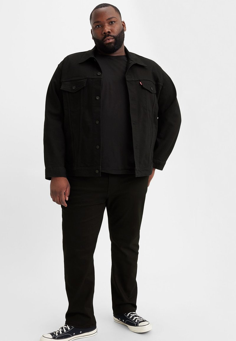 Veste en denim noire, t-shirt noir et jean noir. Modèle portant des baskets bleu marine, le t-shirt a un col rond et la veste possède des boutons-pression.