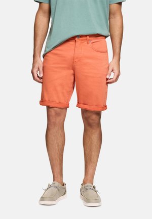 Hombre con pantalones cortos naranjas remangados, una camiseta verde claro y zapatos informales beige, de pie frente a un fondo liso.