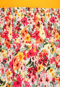 O tecido do vestido floral apresenta um padrão vibrante com vermelhos, laranjas e amarelos contra um fundo claro, com pregas franzidas e uma faixa amarela com nervuras na cintura.