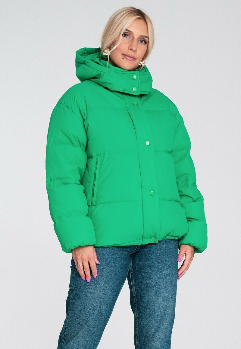 Zielona kurtka pikowana z wysokim kołnierzem, zatrzaskami i szerokimi rękawami. Posiada pikowany wzór i boczne kieszenie. Stylizowana z dżinsowymi spodniami.