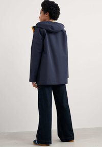 Tamsiai mėlyna vandeniui atspari striukė su kapišonu, turinti geltoną vidinę pamušalą, derinama su plačiomis tamsiai džinsinėmis kelnėmis ir mėlynais šlepetėmis.