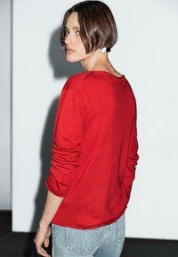 Eine Frau mit kurzen Haaren schaut über ihre Schulter zurück. Sie trägt einen roten Pullover und hellblaue Jeans, vor einem minimalistischen Hintergrund.