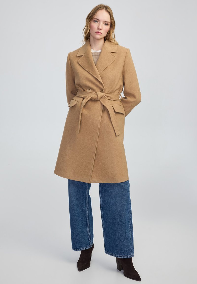 Manteau en mélange de laine couleur camel avec une taille ceinturée, des boutons double boutonnage et des revers. Associé à un jean en denim à jambes larges et des bottes à talons.