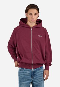 Young man wearing a maroon zip-up hoodie with a hood, front pockets, and light blue jeans, standing against a plain white background.