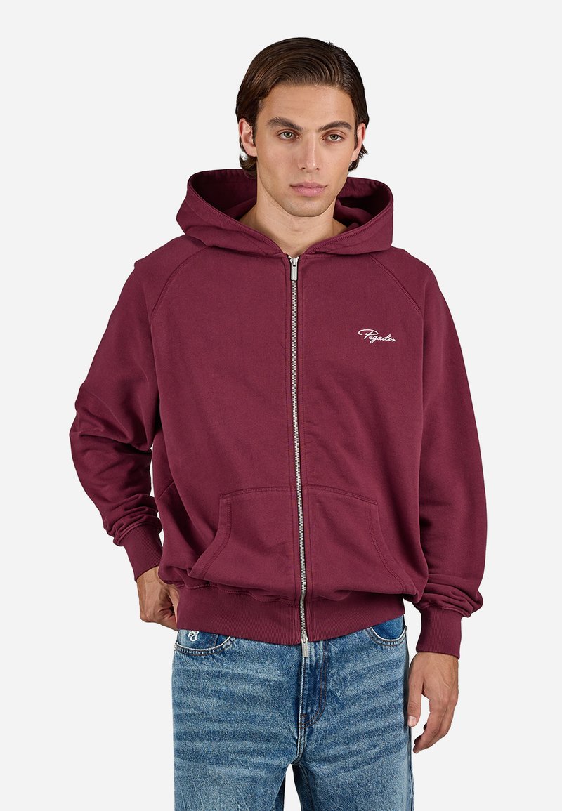 Ung mann iført en bordeaux farget hettejakke med glidelås, frontlommer og lyseblå jeans, som står mot en ensfarget hvit bakgrunn.