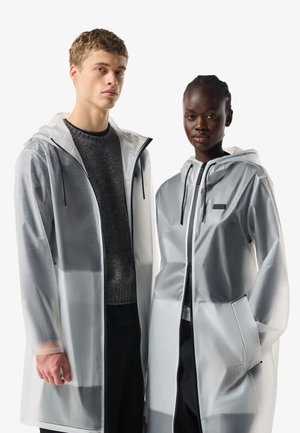 Két modell áttetsző kapucnis esőkabátot visel réteges sötét ruházat fölött, egymás mellett állnak egy egyszínű fehér háttér előtt.