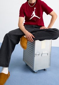 Czerwona koszulka z białym logo Jordana, czarne jeansy o szerokich nogawkach oraz brązowe zamszowe chodaki na srebrnej walizce na kółkach z teksturowaną powierzchnią.