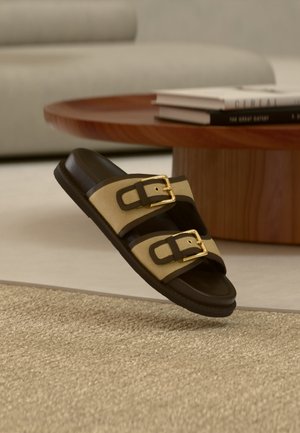 Sandale tip șliț maro și bej cu două catarame aurii, așezate pe un covor bej țesut, lângă o masă rotundă din lemn cu cărți stivuite.