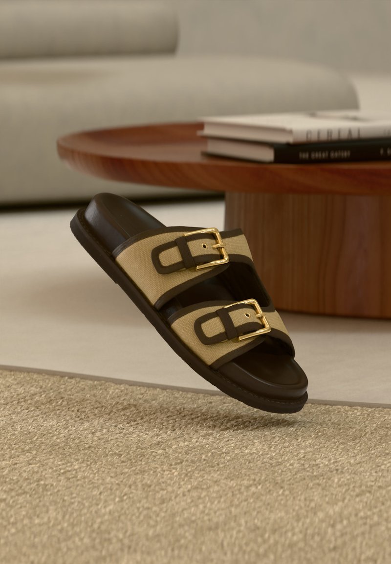 Καφέ και μπεζ δερμάτινο σανδάλι με δύο χρυσά αγκράφες, ακουμπισμένο σε μπεζ υφαντό χαλί κοντά σε στρογγυλό ξύλινο τραπέζι με στοιβαγμένα βιβλία.