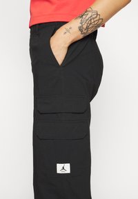 Zwarte cargopants van een textuurstof, met twee grote voorzakken en een witte Jordan-logopatch aan de zijkant. Inclusief elastische tailleband.