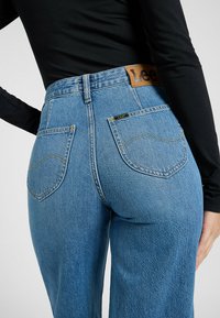 Høyt liv blå denimjeans med kontrasterende gul søm, baklommer og en brun lærlabel på livet.