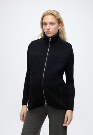 Blonde Frau trägt eine schwarze Fleeceweste mit Reißverschluss über einem schwarzen Langarmshirt und grauen gerippten Hosen vor einem hellen einfarbigen Hintergrund.