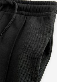 Black jogger pants with a textured fabric, elastic waistband, and adjustable drawstring. Features side pockets and a relaxed fit.