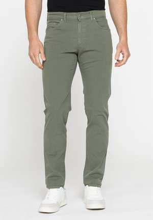 Uomo con pantaloni verde oliva slim fit e sneakers bianche che sta in piedi davanti a uno sfondo chiaro e semplice.