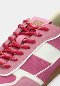 Gros plan sur une basket en daim rose avec des accents blancs, des lacets rouges à motifs et une semelle gomme, mettant en avant les détails des lacets et de la languette.