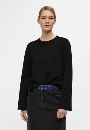 Schwarzer Pullover mit langen Ärmeln und Rundhalsausschnitt, kombiniert mit einem dunkelgrauen Denimrock und einem blauen Gürtel. Glatte Textur, lockerer Schnitt.