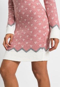 Abito maglione di colore rosa chiaro con motivi bianchi e orlo a zigzag grigio. Presenta polsini a coste bianchi. Tessuto morbido e strutturato.
