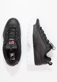 Zapatillas deportivas negras con una parte superior de cuero perforado, suela de goma blanca y cordones negros; presenta un logo y acentos en rojo y blanco.